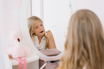 Obraz na płótnie Canvas Child girl paints her lips with glitter or balm in front of a mirror in her room