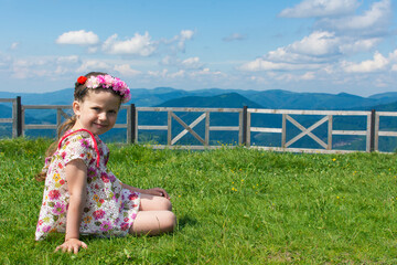 A smiling caucasian girl sits with a wreath on her head on a grass high in the mountains on a summer sunny day in Ukraine.