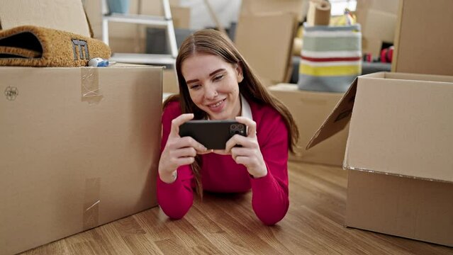 Young hispanic woman watching video on smartphone lying on floor at new home