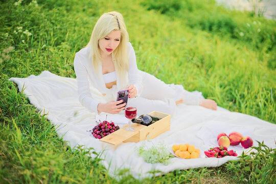 Woman taking pictures of food, wine, cherries, bottle, fruit on her smartphone at a picnic