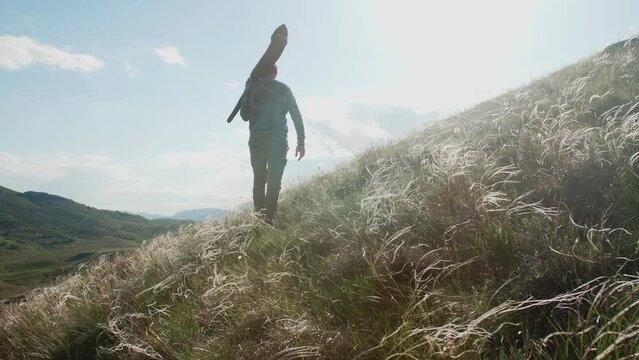 A man walks along a hillside with a feather-grass and carries a Didgeridoo on his shoulder. View from bottom to top. Bright sunny day. The camera is moving