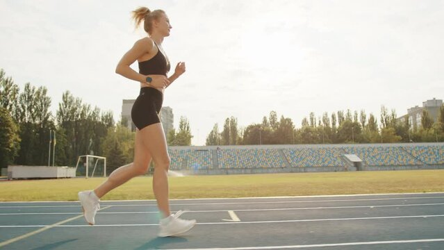 Slim fit young blonde running on athletics track in the stadium on a sunny day. Profile shot of athletic woman performing running workout outdoors. High quality 4k footage