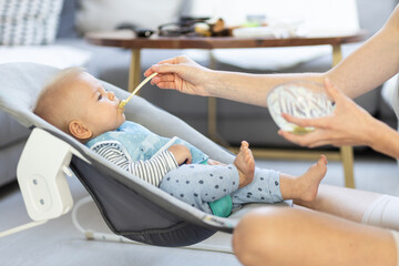 Obraz na płótnie Canvas Mother spoon feeding her baby boy child in baby chair with fruit puree. Baby solid food introduction concept
