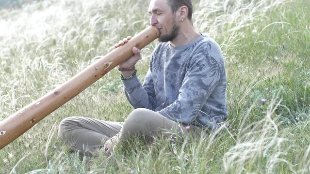Slow motion side view of a hipster caucasian man playing the Australian aboriginal wind musical instrument didgeridoo against the backdrop of green grass. Good musical footage