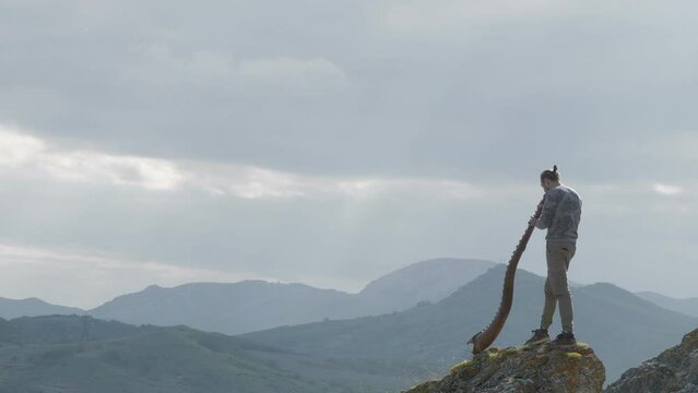 The musician plays the Didgeridoo an Australian aboriginal musical instrument. A man stands on a ledge of a rock on a hillside against the backdrop of a majestic mountain landscape and a cloudy sky