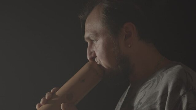 A hipster man plays the Australian aboriginal wind musical instrument Didgeridoo on a dark background. Music footage
