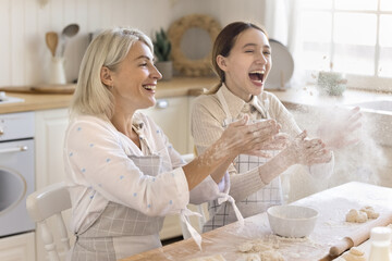 Happy excited teen granddaughter and grandma in aprons having sun, baking in kitchen, throwing...
