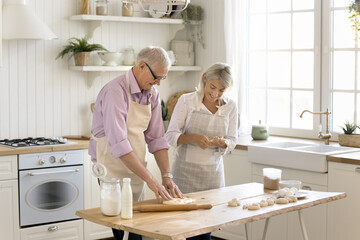Happy mature senior family couple baking together, rolling dough on kitchen table, talking,...