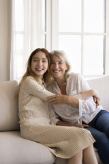 Happy pretty teenager girl embracing cheerful blonde grandmother on home sofa, looking at camera, smiling, laughing, posing for family portrait. Granddaughter and grandma vertical shot