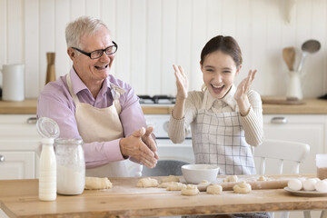 Happy elder grandpa and teenage granddaughter kid baking together in home kitchen, having fun, clapping floury hands over kitchen table with raw dough, laughing, enjoying domestic activities