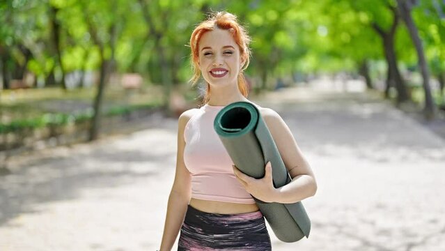 Young redhead woman smiling wearing sportswear holding yoga mat at park