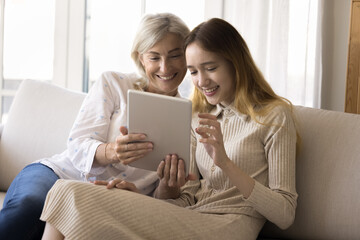 Cheerful teen grandchild girl and happy grandmother holding tablet, using wireless Internet technology at home, enjoying communication, smiling, watching content, shopping online