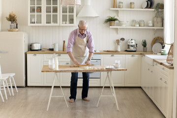 Mature older baker man in apron rolling dough on table with flour, baking ingredients, enjoying culinary cooking activity, preparing dessert for dinner in home kitchen interior