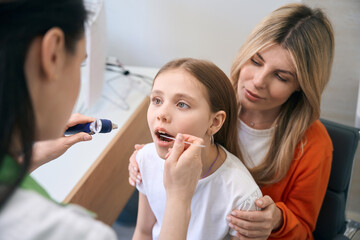 Obraz na płótnie Canvas Laryngologist examining girl throat using sepcial stick and light