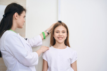 Woman nurse checking little girl height using special measuring plank - obrazy, fototapety, plakaty