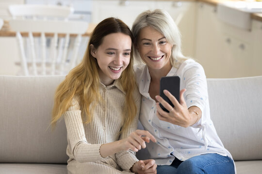 Happy teen grandkid and pretty blonde grandmother taking self picture on smartphone, having fun, smiling, laughing, posing for selfie on mobile phone, enjoying family leisure, friendship