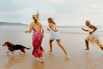 three young girls run along the beach along with a dog. multiracial (caucasian, african and latin) group of female friends enjoying a summer day.