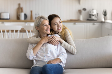 Fototapeta na wymiar Positive dreamy teenage kid hugging mature grandma from behind at home, looking away, smiling, thinking, dreaming, discussing future plans, enjoying family relationships, bonding, affection