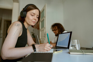Woman in headphones making notes on husband background while they working from home office