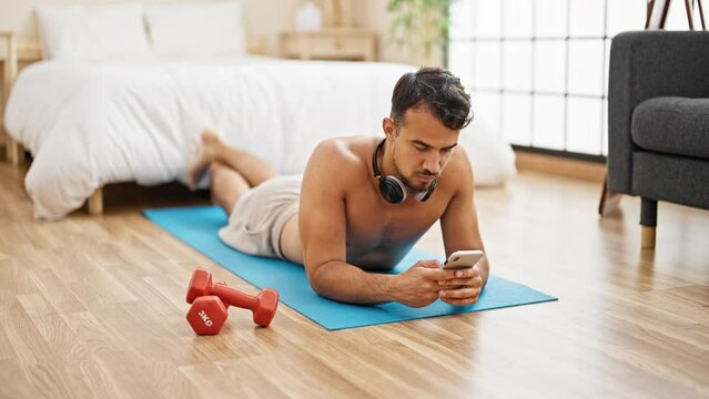 Young hispanic man using smartphone lying on yoga mat at bedroom