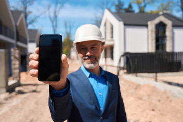 Smiling foreman in hardhat holding and showing smartphone display