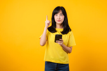 Capture young Asian woman wearing a yellow t-shirt and denim jeans points with finger hand gesture to free copy space while using smartphone. Perfect for illustrating the concept of app functionality.