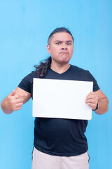 A biracial male with undercut and pursed lips angrily holding a blank placard while pointing at it with one hand. Isolated on a blue background. - obrazy, fototapety, plakaty