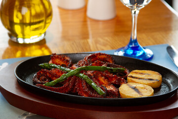 Grilled octopus tentacle served with potatoes and baked asparagus in a clay and wood dish on a table with a glass of wine, oil, salt and pepper. Appetizing dish. Food concept