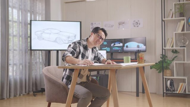 Asian Male Having A Backache While Working On A Car Design Sketch On Table In The Studio With Tv And Computers Display 3D Electric Car Model 
