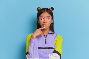 Young Thai woman in sports jacket stands on blue background, holding her hand under chin, thinking, making decision concept, copy space