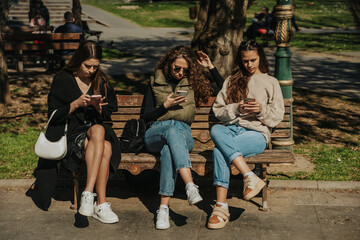 Obraz na płótnie Canvas Three female friends sitting on a bench and not talking. They are scrolling on their social medias