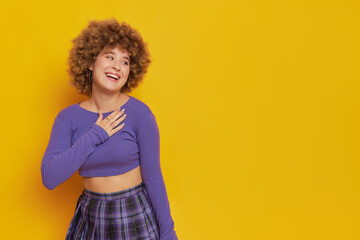 Cute girl with afro hairstyle dressed in purple longsleeve top on yellow background, satisfied smile, hands pressed to heart, happy moments concept, copy space