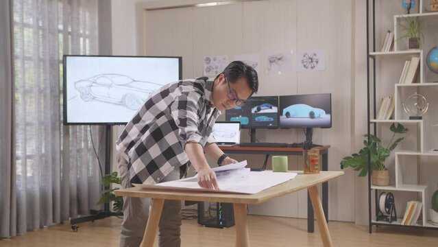 Asian Male Organizing Papers While Working About Car Design Sketch On Table In The Studio With Tv And Computers Display 3D Electric Car Model 
