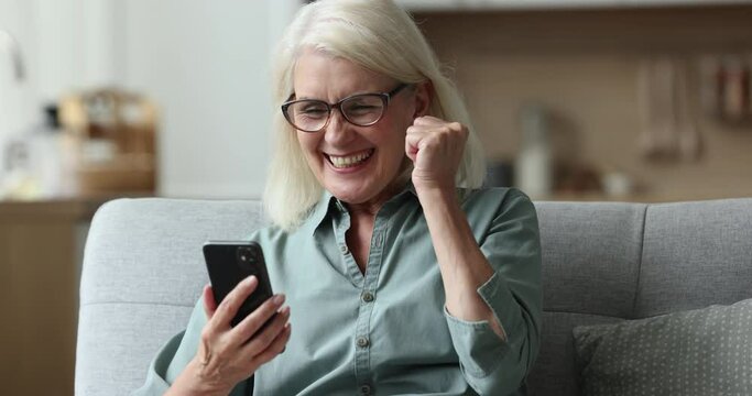 Serious focused senior blonde woman in glasses, using Internet technology on mobile phone, resting on couch at home, getting surprised with good news, celebrating achievement, making winner hand