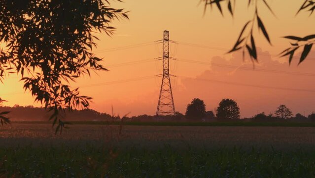 Silhouette of high voltage pole next to road with moving cars at sunset, view from agricultural field. Electricity price concept.