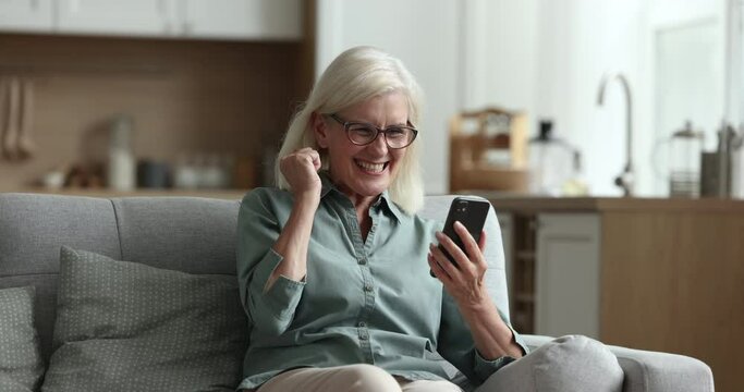 Positive old mature blonde lady in eyeglasses, holding mobile phone, relaxing on home sofa, enjoying leisure, browsing Internet on smartphone, getting cheerful, happy, shouting yes with win hand