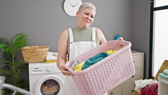 Middle age grey-haired woman smiling confident holding basket with clothes at laundry room