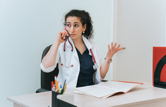 Annoyed Brunette Female Doctor Sitting At Desk Talks By Phone With Unpleasant Face Expression. Nurse Arguing With Patient  Explains Necessity Of Treatment. Puzzled Doctor Working.