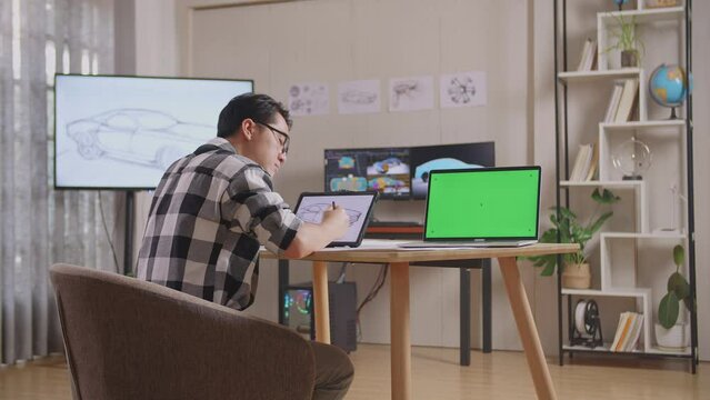 Side View Of Asian Male Thinking About Car Concept On The Table With A Tablet Beside Green Screen Laptop In The Studio With Tv And Computers Display 3D Electric Car Model 
