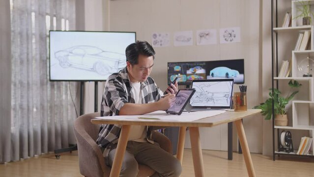Side View Of Asian Male Looking At Smartphone And Drawing New Car Design Concept On A Tablet In The Studio With Tv And Computers Display 3D Electric Car Model 
