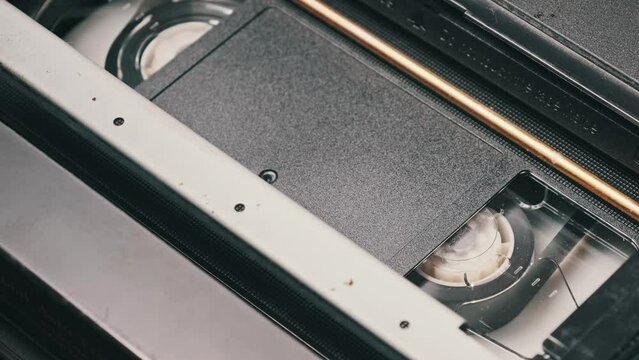 VHS cassette is played inside a VCR tape recorder, top view. Reels of videotape rotate. Video cassette with a blank tag is starting playback. Old video recorder inside close-up. Playing old movie.