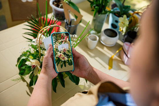 Close-up female florist holding a mobile phone and photographing a bouquet of flowers in wicker basket, created in floral design studio. Smartphone in live view mode. Wedding, Mother's and Women's Day