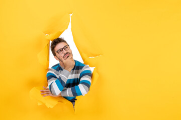 front view thinking young man putting hand on his chin peeping through hole in paper yellow wall