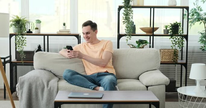 Young Caucasian man sits comfortably on a couch, radiating happiness. With a warm smile on his face, he effortlessly navigates his smartphone, fully engaged in the digital world.