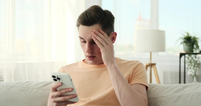 Man sitting on a sofa in the living room, with a look of disappointment on his face. He holds a phone in his hands, which he appears to be using, although the exact nature of his activity is unclear.