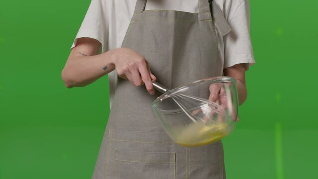 Close-up of Caucasian woman beats fresh eggs into the glass bowl and puts egg shells to the small dish on green screen background. Female stands in her kitchen