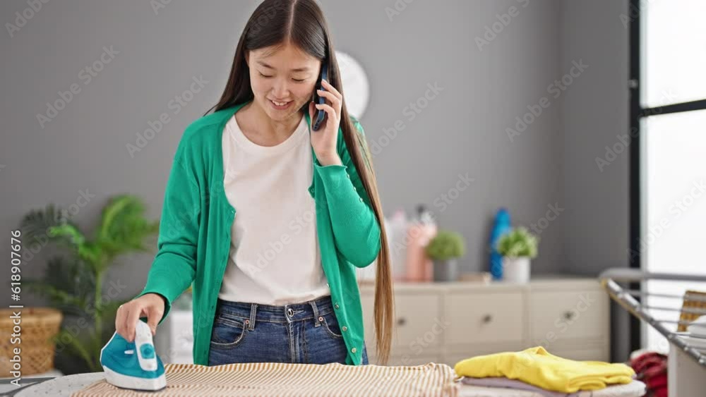 Wall mural Young chinese woman talking on smartphone ironing clothes at laundry room - Wall murals