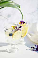 Iced soda with lemon and edible flowers under palm leaves on a bright sunny day. Summer cold cocktails and drinks. Detox water.