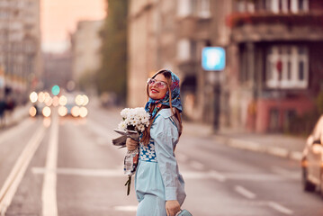 Charm of a woman adorned in an exquisite blue traditional dress, carrying a blue handbag and a bouquet of flowers, gracefully strolling through the city at sunset, creating a mesmerizing scene of