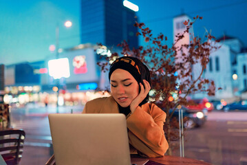 In the enchanting ambiance of a nocturnal cityscape, a hijab-clad girl engrossed in her laptop...